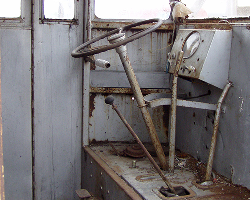 This is a view of the interior of the SF Divco. In addition to the steering wheel column, there is a gear shift, then a hand brake to slow down while still driving and going around corners and the emergency brake handle.